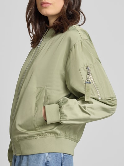 Frauenmodel trägt Regular Fit Bomberjacke mit College-Kragen von s.Oliver RED LABEL w kolorze jednolitym, satynowa powierzchnia, zamek błyskawiczny, zawieszka z logo, długi rękaw z prążkowanym wykończeniem, wpuszczane kieszenie i brustasza, widok na górę z przodu.