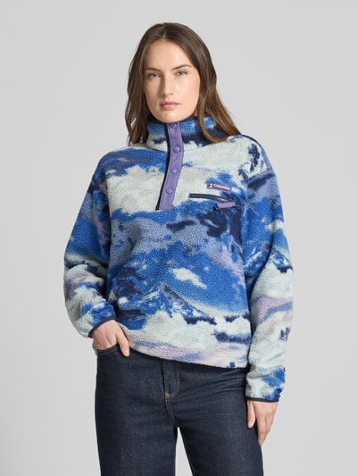 Frauenmodel trägt Regular Fit Pullover mit Logo-Patch Modell 'Helvetia' von Columbia in Fleece-Optik, blau-gemustert, Knopfleiste, Stehkragen, Brusttasche, elastische Ärmelabschlüsse, Ganzkörperansicht von vorne.
