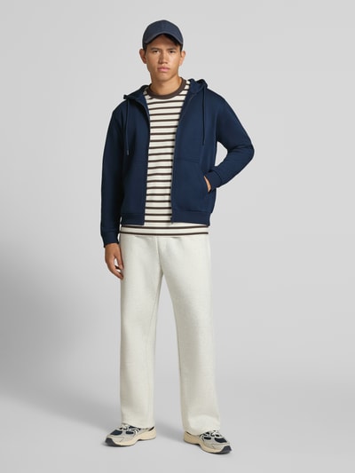 Männermodel trägt Regular Fit Sweatjacke aus Baumwoll-Mix Modell 'BRADLEY' von JACK & JONES in Dunkelblau, Ganzkörperansicht von vorne, Herren Sweatjacke mit Kapuze, Tunnelzug, Känguru-Tasche, elastische Abschlüsse, Rückenlänge 67 cm, Ärmellänge 68 cm.