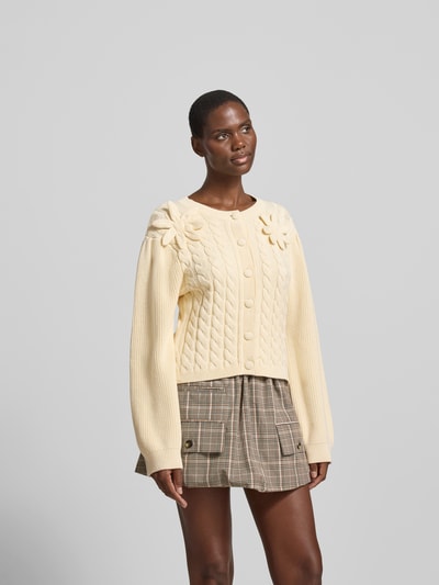 Frauenmodel trägt Strickjacke mit floralem Zierbesatz von BAUM & PFERDGARTEN, Reine Baumwolle, taillierte Passform, Rundhalsausschnitt, durchgängige Knopfleiste, Rückenlänge bei Größe S 53 cm, cremefarben mit Strukturmuster, Ganzkörperansicht von vorne.