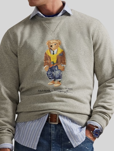 Męski model nosi Regular Fit Sweatshirt aus Baumwoll-Mix mit Polo Bear od Polo Ralph Lauren, melanż, krój regular fit, aplikacja nadruk Polo Bear, okrągły dekolt, długi rękaw, zakończenia prążkowane, dzianina dresowa, mieszanka bawełny, zbliżenie na górną część ciała.