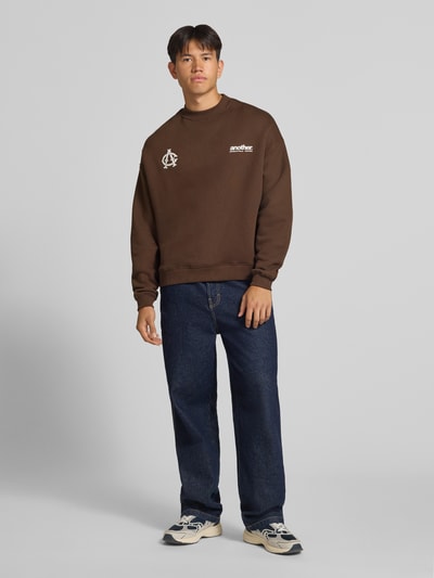 Męski model nosi brązową bluzę 'Sweatshirt mit Label-Stickerei' od ANOTHER COTTON LAB, mieszanka bawełny, luźny krój, okrągły dekolt, wyhaftowane logo, prążkowane wykończenia, długość tyłu w rozmiarze M 63 cm, długość rękawów w rozmiarze M 58 cm, widok całej sylwetki z przodu.