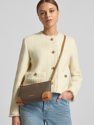 Frauenmodel trägt Handtasche mit Allover-Logo-Print Modell 'Murphy' von MICHAEL Michael Kors, braune und beigen Farbtöne, strukturierte Oberfläche, schmaler Schulterriemen, Reißverschlussfach, Ganzkörperansicht von vorne.