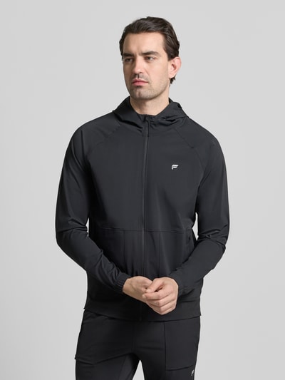 Mannenmodel draagt trainingsjack met capuchon THE ONE JACKET van Fabletics in zwart, reflecterend logo, elastische mix, losse fit, doorlopende ritssluiting, opgestikte ritszakken, elastische mouwboorden. Vooraanzicht bovenlichaam.