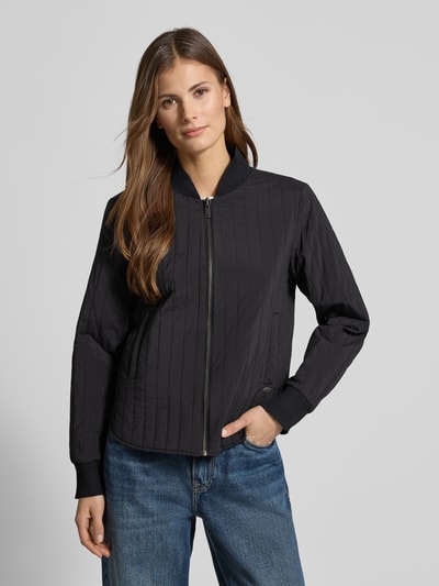 Vrouwenmodel draagt Jacke met Stehkragen und Eingrifftaschen van Scotch & Soda, regular fit, zwart met labeldetail, ritssluiting, steekzakken, ruglengte maat S 57 cm, schouderbreedte 44 cm, borstwijdte 50 cm, mouwlengte 63 cm, ganzkörperansicht van voren.