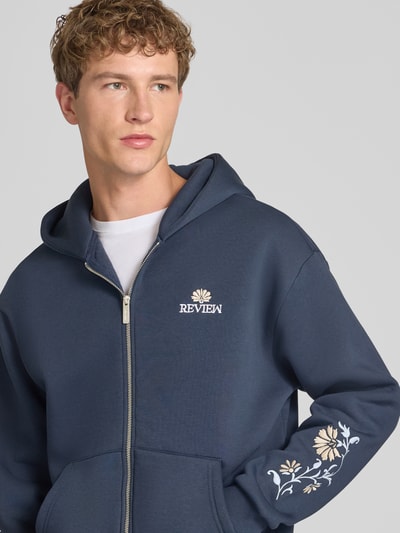 Männermodel trägt Relaxed Fit Sweatjacke mit Kapuze von REVIEW in Dunkelblau mit floraler Stickerei, Nahaufnahme Oberkörper. Baumwoll-Mix, Kängurutasche, Reißverschluss, Rückenlänge 60 cm, Ärmellänge 63 cm.