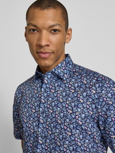 Mężczyzna model nosi Regular Fit Freizeithemd mit Kentkragen od OLYMP, krój regular fit, florystyczny nadruk allover, krótki rękaw, pełna listwa guzikowa, długość pleców w rozmiarze 40: 85 cm, szerokość w rozmiarze 40: 45 cm, długość rękawa w rozmiarze 40: 28 cm, granatowe tło z kolorowymi kwiatami, tkanina gładka, zbliżenie na górną część ciała, widok z przodu.