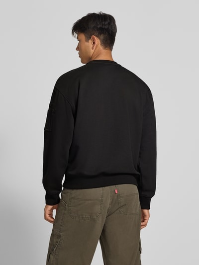 Męski model nosi bluzę Sweatshirt mit Label-Details von Alpha Industries, mieszanka bawełny, luźny krój, okrągły dekolt, aplikacja z metką na kieszeni na rękawie, prążkowane wykończenia, kolor czarny, zdjęcie całej sylwetki z tyłu.