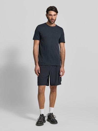 Męski model nosi Slim Fit T-Shirt mit Logo-Print Modell 'Vanish' od Under Armour, czarny kolor, okrągły dekolt, dziurkowana struktura, szerokość w rozmiarze M 40 cm, długość nogawki wewnętrznej 70 cm, krój slim fit, widok całej sylwetki z przodu.