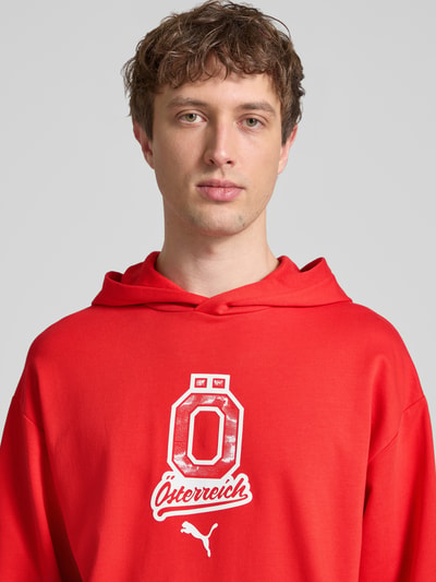 Männermodel trägt Relaxed Fit Hoodie mit Känguru-Tasche von PUMA PERFORMANCE in kräftigem Rot mit Label-Detail, Nahaufnahme Oberkörper.