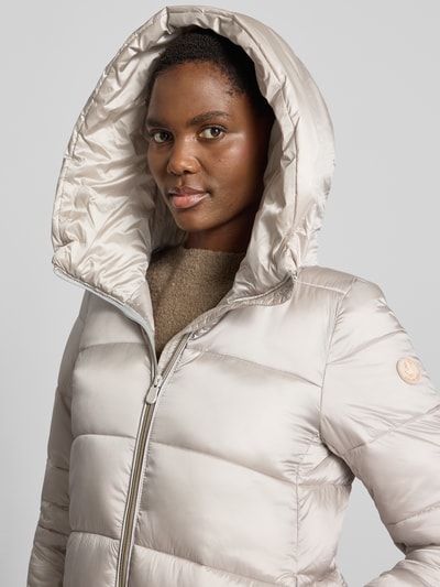 Frauenmodel trägt Steppmantel mit Kapuze Modell 'LYSA' von Save The Duck in Beige, glatte gesteppt-glänzende Oberfläche, Label-Patch, Nahaufnahme Oberkörper. Kontext: Gerader Schnitt, Zweiwege-Reißverschluss, seitliche Eingrifftaschen, Rückenlänge 91 cm, Schulterbreite 39 cm, Brustweite 40 cm, Ärmellänge 66 cm.