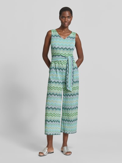 Frauenmodel trägt Jumpsuit mit Bindegürtel und V-Ausschnitt von Betty Barclay im Zickzack-Muster, Material-Mix mit Stretch-Anteil, Taillierter Schnitt, Rückseitiger Reißverschluss, Creme-Weiß-Grün-Blau, Rückenlänge 34 cm, Brustweite 44 cm, Gesamtlänge 132 cm, Innenbeinlänge 63 cm, Ganzkörperansicht von vorne.