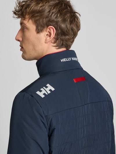 Männermodel trägt Regular Fit Jacke mit Stehkragen von Helly Hansen in Dunkelblau mit Logo-Print, Eingrifftaschen, Reißverschluss, Nahaufnahme Rückansicht. Rückenlänge 70 cm, Schulterbreite 46 cm, Brustweite 54 cm, Ärmellänge 69 cm, Größe M.