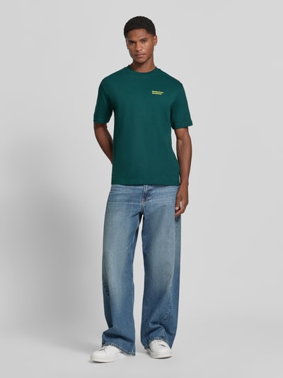 Männermodel trägt T-Shirt mit Statement-Print von MCNEAL in Dunkelgrün, lockerer Schnitt, kombiniert mit Jeans, Ganzkörperansicht von vorne. Rückenlänge bei Größe M: 66 cm, Rundhalsausschnitt.