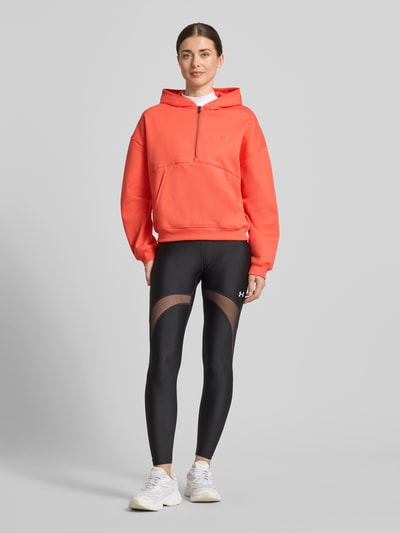 Modelka kobieca ma na sobie Hoodie mit Reißverschluss und Label-Stitching od Under Armour, kolor koralowy, bawełna, krój regular fit, z kapturem, kieszenią kangurką, widok całej sylwetki z przodu.
