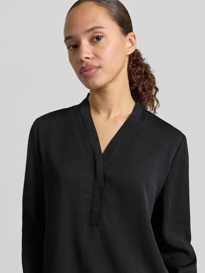 Frauenmodel trägt Bluse mit V-Ausschnitt und Label-Detail von Marc Cain in Schwarz, Loose-Fit, elastische Ärmelabschlüsse, Nahaufnahme Oberkörper, Rückenlänge 46 cm, Ärmellänge 60 cm, Größe 36.