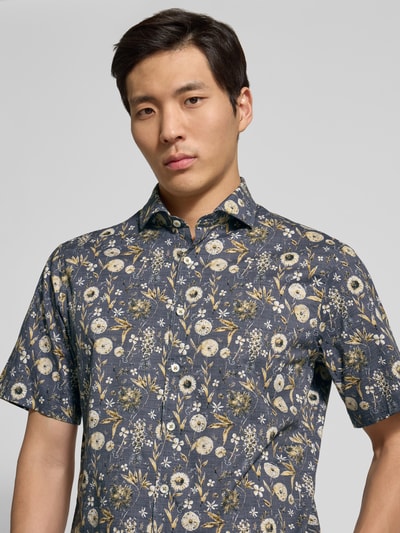 Męski model nosi Modern Fit Freizeithemd im Allover-Look Modell 'Hardy' od Brax, ciemny wzór, krótki rękaw, koszula, półzbliżenie górnej części ciała, Kentkragen, zapięcie na guziki, długość pleców w rozmiarze M: 76 cm, szerokość ramion w rozmiarze M: 48 cm, długość rękawa w rozmiarze M: 25 cm.