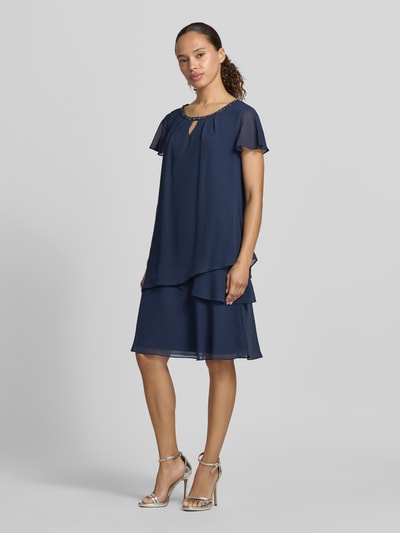 Frauenmodel trägt knielanges Cocktailkleid mit Schlüsselloch-Ausschnitt von Christian Berg Cocktail in Dunkelblau, Chiffon, Stufen-Look, Perlen, Gerader-Schnitt, Ganzkörperansicht von vorne.
