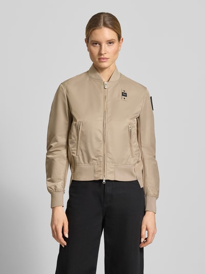 Frauenmodel trägt Bomberjacke mit Reißverschlusstaschen Modell 'LAURIE' von Blauer Usa in Beige mit Stehkragen, Zweiwege-Reißverschluss, Label-Patches und gerippte Abschlüsse, Loose-Fit, seitliche Reißverschlusstaschen; glatte Textur, Kontrast durch schwarze Hose, Ganzkörperansicht von vorne.