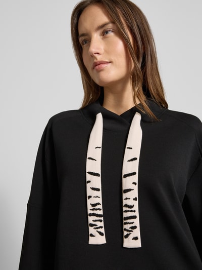 Frauenmodel trägt Oversized Sweatshirt mit Tunnelzug von Marc Cain aus Baumwoll-Mix mit Stretch-Anteil, Schwarz mit Kontrastdetails, Nahaufnahme Oberkörper.