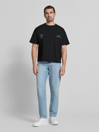Męski model nosi Regular Fit T-Shirt mit überschnittenen Schultern von JACK & JONES, mieszanka bawełny, czarny, okrągły dekolt, nadruk z napisem na tyle, długość tyłu w rozmiarze M: 66 cm, ganzkörperansicht von vorne