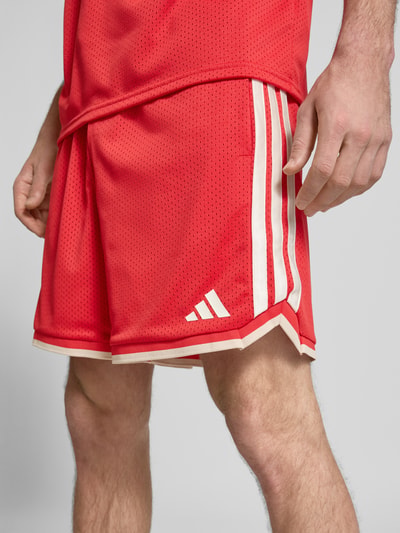 Männermodel trägt Shorts mit elastischem Bund und Kordelzug von adidas Sportswear in Rot mit weißen Streifen, Nahaufnahme Oberschenkel. Lockere Passform, Logo-Print, sichtbare Struktur des Stoffs.
