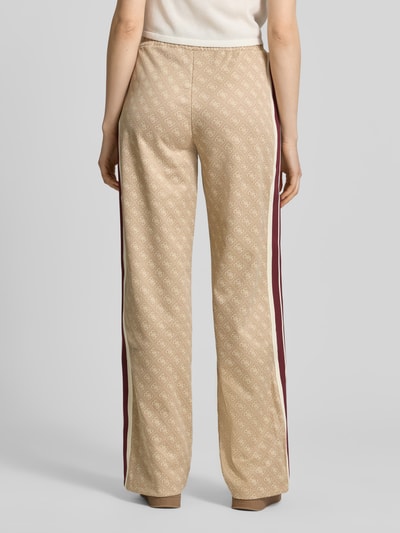 Frauenmodel trägt Wide Fit Sweatpants mit Allover-Logo-Print Modell 'Jacquard' von Guess Activewear in Beige mit Kontraststreifen, Material-Mix mit Baumwoll-Anteil, elastischer Bund, Eingrifftaschen, Ganzkörperansicht Rückansicht.