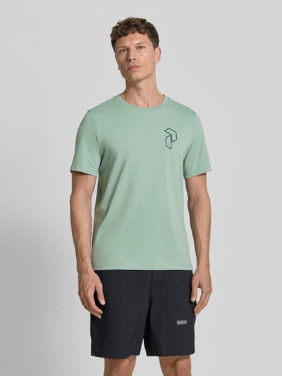 Männermodel trägt T-Shirt mit Label-Print und Rundhalsausschnitt von PEAK PERFORMANCE in Mintgrün, Regular Fit, Ganzkörperansicht von vorne. Reine Baumwolle, dezentes Logo links, kombiniert mit schwarzen Shorts. Brustweite 54 cm, Schulterbreite 46 cm, Ärmellänge 25 cm, Rückenlänge 71 cm bei Größe M.