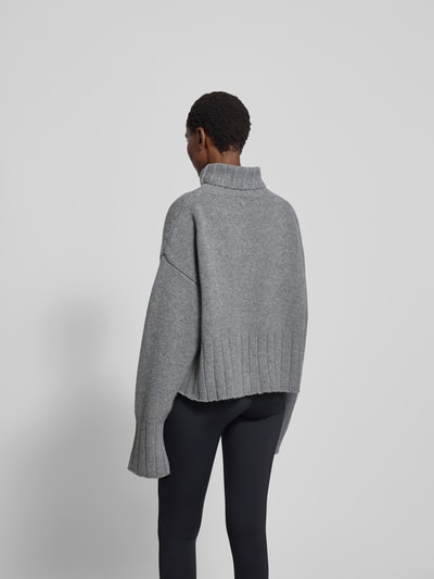 Frauenmodel trägt Oversized Pullover aus reiner Wolle von black palms in Grau mit gerippten Abschlüssen und Rollkragen, Rückenlänge 56 cm, Ganzkörperansicht Rückansicht.