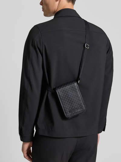 Männermodel trägt Handytasche mit Allover-Muster Modell 'EMBLEM AOP' von CK Calvin Klein für Herren in dunklen Tönen, Ganzkörperansicht Rückansicht.
