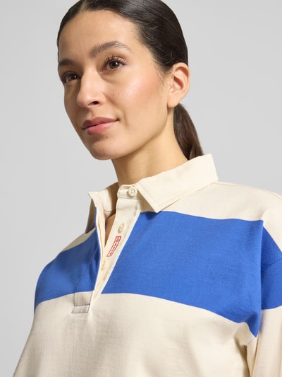 Frauenmodel trägt Regular Fit Langarm-Poloshirt mit verdeckter Knopfleiste von Gant aus reiner Baumwolle, creme mit blauer Bruststreifen, Nahaufnahme Oberkörper.