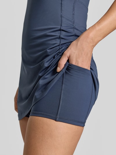 Kobiece model prezentuje sukienkę mini z zintegrowanymi szortami od VARLEY, elastyczna mieszanka materiałów, krój slim fit, kieszeń w pasie, bez rękawów, długość tyłu w rozmiarze S: 79 cm, szerokość w rozmiarze S: 33 cm, niebieski kolor, gładka tekstura, detaliczny widok boczny na biodro