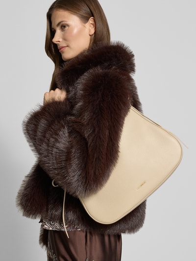 Frauenmodel trägt Handtasche mit Label-Detail Modell 'NORY' von Coccinelle, beige, Hauptfach mit Reißverschluss, abnehmbarer Tragehenkel, Innenfach mit Reißverschluss, Breite 28 cm, Länge 26 cm, Tiefe 9 cm, Schulterriemen 109 cm, Ganzkörperansicht von vorne.