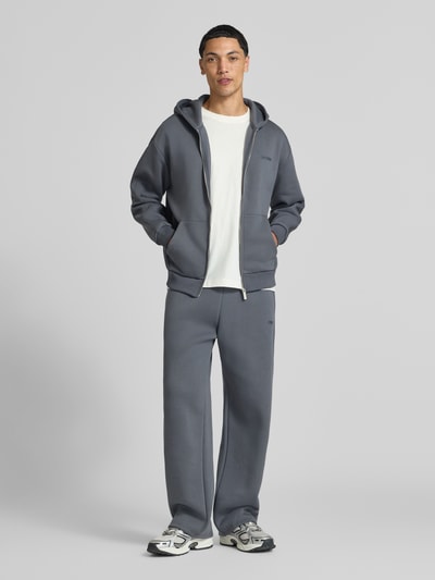 Männermodel trägt Relaxed Fit Sweatjacke aus Baumwoll-Mix mit Label-Detail von REVIEW, gedeckte Grautöne, glatte Textur, Ganzkörperansicht von vorne. Kontext: Herren Sweatjacke, Kapuze, Reißverschluss, geteilte Känguru-Tasche, elastische Abschlüsse.