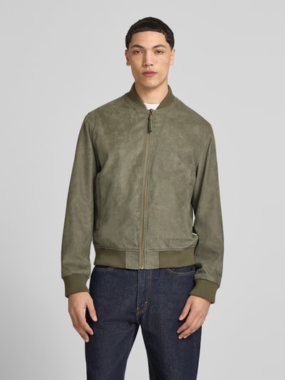 Męski model nosi bomberka in Leder-Optik mit Stehkragen von Jack & Jones, krój regular fit, wpuszczane kieszenie, zamek błyskawiczny, długość tyłu w rozmiarze M 64 cm, szerokość ramion w rozmiarze M 45 cm, szerokość klatki piersiowej w rozmiarze M 52 cm, długość rękawów w rozmiarze M 67 cm, oliwkowa, gładka faktura, ciemnoniebieskie jeansy, całkowity widok z przodu.