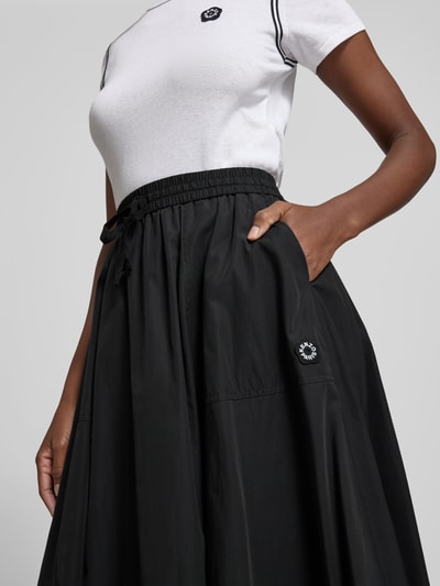 Frauenmodel trägt Midirock mit Baumwoll-Anteil von Kenzo in Schwarz, A-Linie, elastischer Bund mit Tunnelzug, Label-Detail, Nahaufnahme Oberkörper.