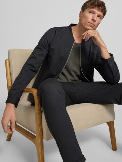 Męski model nosi Regular Fit Sweatjacke mit Stehkragen od Lindbergh, elastyczna mieszanka materiałów, wpuszczane kieszenie, zamek błyskawiczny, długość tyłu w rozmiarze M 69 cm, długość rękawów 66 cm, kolor czarny, gładka tekstura, krój regular fit, całkowity widok z przodu.