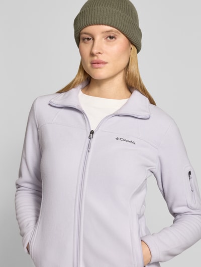 Frauenmodel trägt Slim Fit Fleecejacke mit Ärmeltasche Modell 'Fast Trek™ II' von Columbia in hellem Violett mit Stehkragen und Reißverschluss, elastische Abschlüsse, Label-Detail, Reißverschlusstaschen, Fleece-Textur, Nahaufnahme Oberkörper, Kontext: Damen Fleecejacke aus Fleece, Rückenlänge 63 cm, Schulterbreite 40 cm, Brustweite 45 cm, Ärmellänge 62 cm.