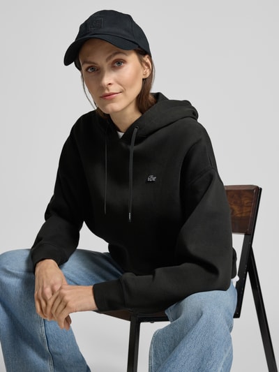 Frauenmodel trägt Oversized Hoodie aus Baumwoll-Mix von Lacoste Sport in Schwarz mit Logo-Detail, Sweatoptik, Kapuze, Nahaufnahme Oberkörper.