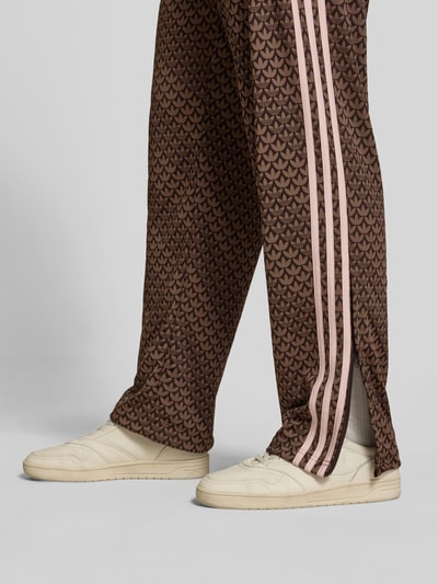 Mężczyzna nosi spodnie dresowe Loose Fit Sweatpants mit Logo-Muster model 'FIREBIRD' von adidas Originals, efekt: wzór z logo na całej powierzchni, krój loose fit, wyhaftowane logo, elastyczny ściągacz z tunelem, boczna kieszeń zapinana na zamek błyskawiczny, wykończenie materiału: dzianina dresowa, detal: jasne paski; zdjęcie: zbliżenie dolnej części z przodu