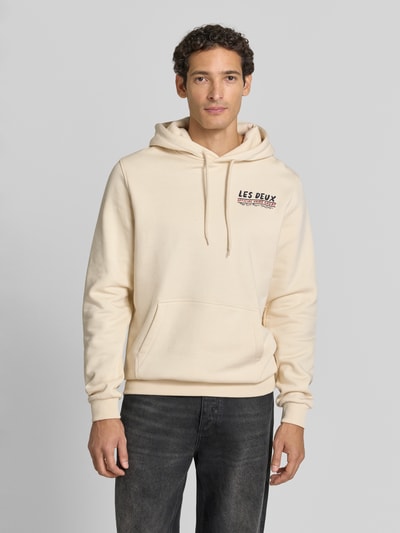 Männermodel trägt Hoodie aus reiner Baumwolle mit Logo-Print Modell 'Record' von Les Deux, helle Farbe, Kängurutasche, Ganzkörperansicht von vorne, Herren Hoodie Regular Fit, Kapuze mit Tunnelzug, gerippte Abschlüsse, Logo-Label, Rückenlänge 70 cm, Ärmellänge 74 cm.