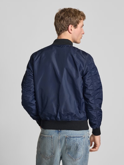 Mężczyzna prezentuje Regular Fit Bomberjacke mit Logo-Patch od Alpha Industries, ciemnoniebieska, gładka faktura, krój regular fit, logo-patch, kieszenie, zamek, kieszeń na rękawie z zamkiem, długość tyłu w rozmiarze M: 65 cm, szerokość ramion w rozmiarze M: 45 cm, szerokość klatki piersiowej w rozmiarze M: 54 cm, długość rękawa w rozmiarze M: 69 cm, całkowity widok z tyłu