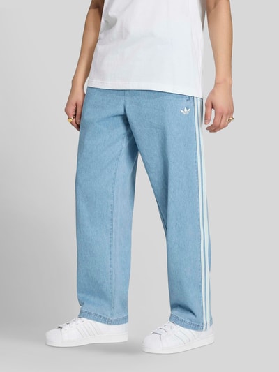 Männermodel trägt Relaxed Fit Trainingshose aus Baumwoll-Mix von adidas Originals in Denim-Optik mit Logo-Applikation, Eingrifftaschen mit Reißverschluss und elastischem Bundabschluss, hellblauer Farbton, Ganzkörperansicht von vorne.