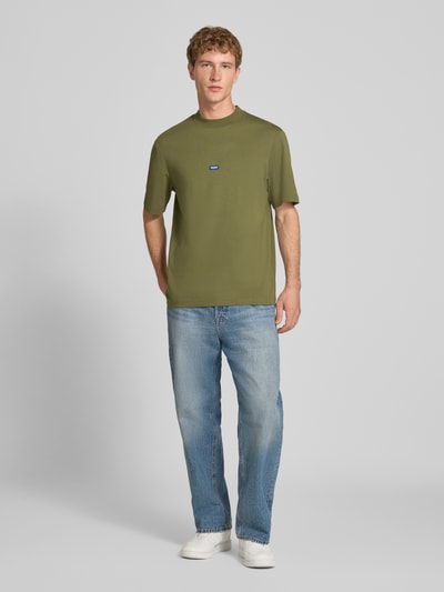 Mężczyzna nosi Męski t-shirt Regular Fit T-Shirt aus reiner Baumwolle model 'NIEROS' od HUGO w kolorze oliwkowym, jasnoniebieskie dżinsy, białe tenisówki, neutralne tło, Ganzkörperansicht von vorne.