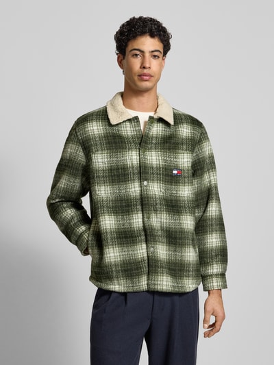 Mężczyzna model nosi Oversized Hemdjacke mit Allover-Muster von Tommy Jeans, efekt: wzór na całej powierzchni, krój oversized, wykładany kołnierz, listwa z zatrzaskami, naszywka z logo, długi rękaw, zakończenia rękawów na zatrzask, wpuszczane kieszenie boczne, kieszenie na piersi, zielone tony, tekstura miękka, Ganzkörperansicht z przodu.