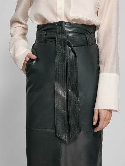 Frauenmodel trägt Eng anliegender Midi-Rock mit seitlichen Eingrifftaschen Modell 'VACARO' von BOSS Orange, Optik: Unifarben, Kunstleder, Label-Stitching-Applikation, Metall-Akzente, Bindegürtel, Gehschlitz, Nahaufnahme Oberkörper.