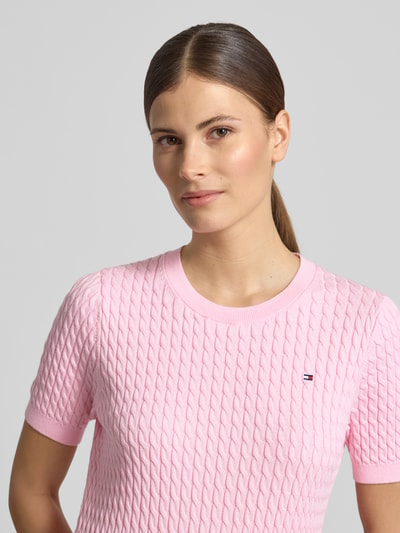 Frauenmodel trägt Slim Fit T-Shirt aus reiner Baumwolle von Tommy Hilfiger in Optik: Unifarben mit Zopfmuster, Kurzarm, Logo-Detail, Strick, Farbe Rosa, Nahaufnahme Oberkörper.