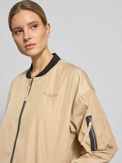 Frauenmodel trägt Relaxed Fit Jacke mit Logo-Stitching von Original Bombers in Beige mit schwarzem Stehkragen, Nahaufnahme Oberkörper. Rückenlänge 84 cm, Schulterbreite 58 cm, Brustweite 56 cm, Ärmellänge 57 cm, Reißverschluss, Eingrifftaschen mit Druckknopf, Armtasche, elastische Abschlüsse.