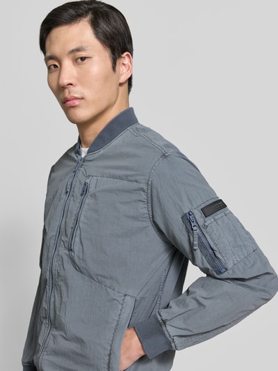 Männermodel trägt khujo Blouson mit Label-Patches Modell 'IBE', Reißverschluss, Brusttasche, Reißverschlusstasche am Ärmel, Seitliche Eingrifftaschen, Gerader-Schnitt, Stehkragen, graue Textur, Nahaufnahme Oberkörper.