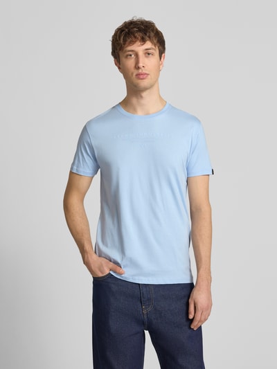 Mannenmodel draagt T-shirt met Label-Print en Rundhalsausschnitt van Alpha Industries in lichtblauw, puur katoen, regular fit, ruglengte maat M 67 cm, voorkant bovenlichaam close-up.